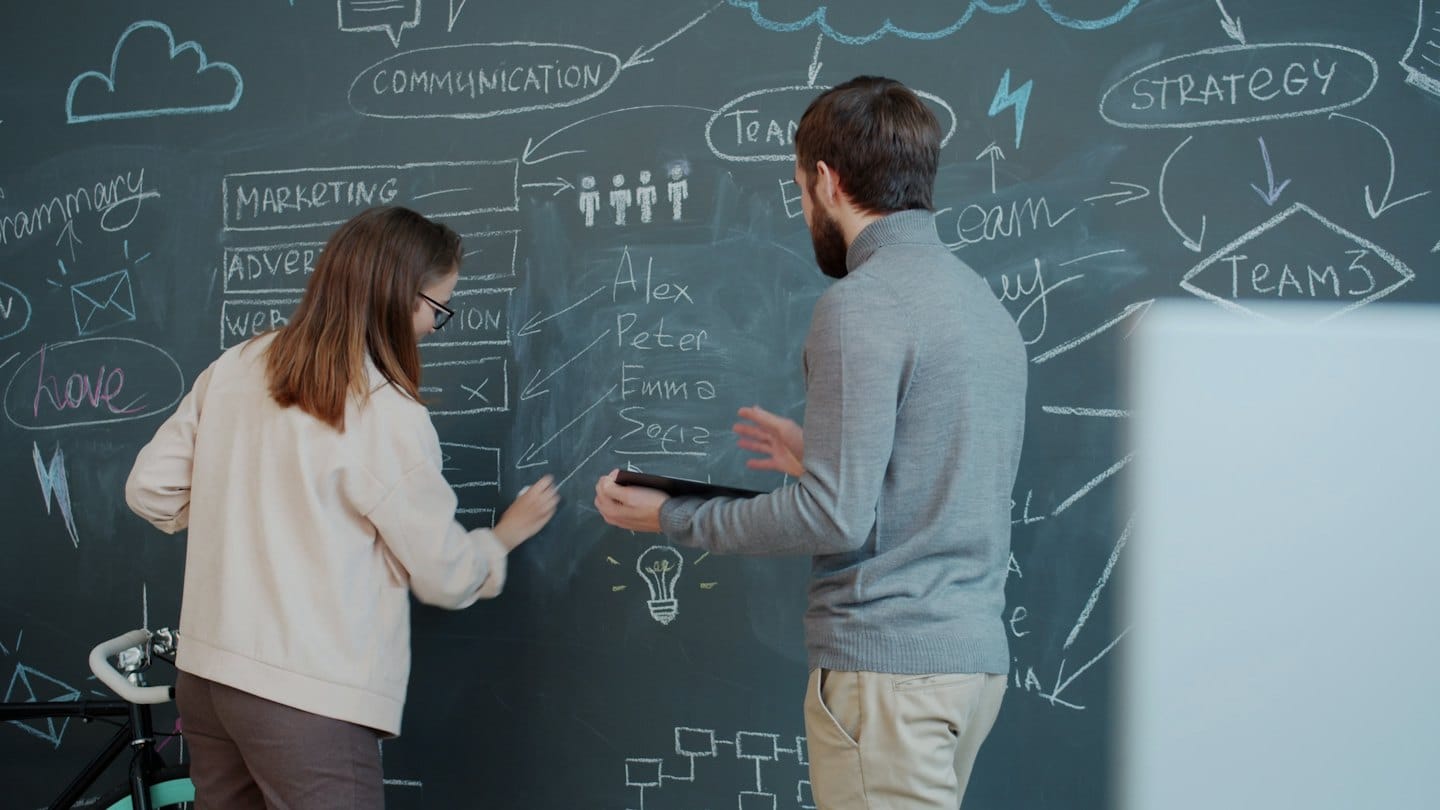 Dvaja kolegovia si vymieňajú nápady na marketingovú stratégiu na veľkej tabuli.